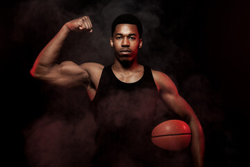 Wall Mural - Basketball player side lit with red color holding a ball against hazy smoke fog background. Serious african american man showing muscles