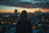 Fototapeta  - A contemplative woman stands with her back to the viewer, watching a stunning sunset over a cityscape of high-rises and lights.