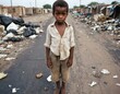 A young african american boy stands on a dirty area in a trash-filled area. He is wearing a dirty and torn clothes