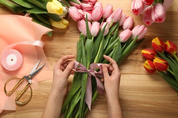 Wall Mural - Woman making beautiful bouquet of fresh tulips and ribbon at wooden table, top view