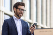 © Tetiana - Close-up photo of a smiling young man businessman, lawyer, politician wearing a suit and glasses standing outside an office building, holding a phone and looking at the screen