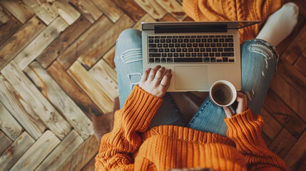 Canvas Print - A person relaxes at home in a cozy orange sweater and denim jeans, using a laptop and holding a coffee mug, exemplifying a comfortable remote work setting.