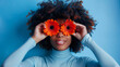 A person holds bright orange gerbera flowers over their eyes like glasses, smiling broadly against a blue background, creating a playful and joyful portrait.