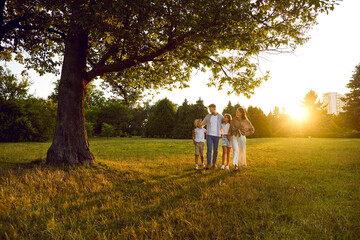 Wall Mural - Family taking stroll on quiet summer evening. Parents and children enjoying beauty of nature. Happy mother, father and kids walking under magnificent green tree on grassy meadow in beautiful city park