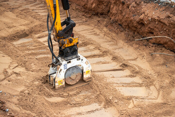 Wall Mural - A mini excavator rams the ground with a vibrating plate. Laying of underground sewer pipes and communications during construction. soil compaction. Earthworks, excavation.