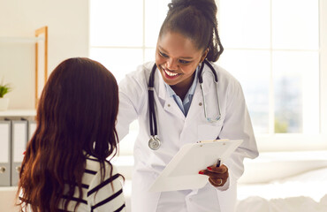 Wall Mural - Friendly dark-skinned female doctor discussing treatment with patient after medical examination. African american doctor with stethoscope and clipboard smiling while touching shoulder of woman.