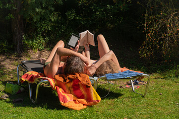 Two young people relax on a sun lounger and read electronic and paper books