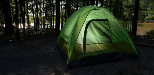 Canvas Print - Shaded campsite in a thick forest with sun on the lake behind and with tent lighted from the interior in the foreground.