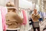 Fototapeta Boho - A young woman tries on a warm jacket in front of a mirror in a store. Cute blonde with short hair. Style and fashion.