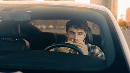 Caucasian man driver businessman eating cheeseburger inside auto in city parking lot. Hungry male guy eat hamburger junk fast food takeaway service in travel enjoying tasty lunch while sit car seat