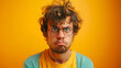 A funny young man with curly hair and glasses looking at the camera, frowning in anger on a yellow background