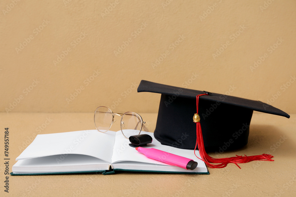 Mortar board with notebook, marker and eyeglasses on brown background