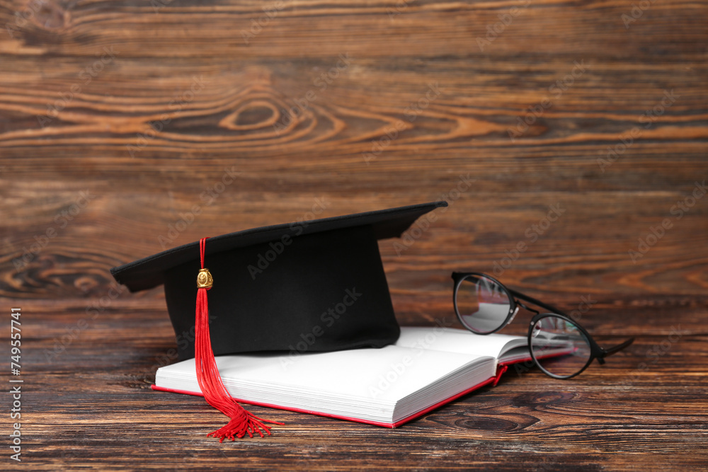 Mortar board with notebook and eyeglasses on wooden background