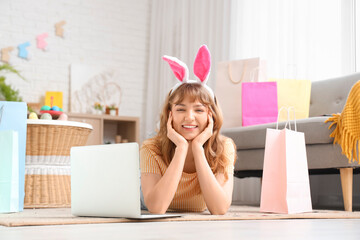 Wall Mural - Happy young woman in bunny ears headband with laptop and paper bags lying on floor at home. Online shopping for Easter concept