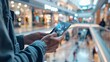 a man's hand holding a mobile smartphone, tablet, and cellphone over a blurred shopping center background.