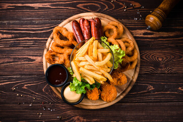 Sticker - Fried chicken legs, crispy onion rings, breaded squid rings, sausages, french fries, sauces and greens on wooden table.