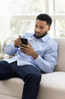 © fizkes - Serious young African business man resting on soft couch in home office, reading, watching online content on smartphone, using financial service, Internet application. Vertical shot