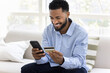© fizkes - Cheerful young African customer man enjoying online shopping, paying for purchase by credit card, using webstore ecommerce service, checking bank account balance
