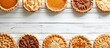 A variety of freshly baked homemade autumn pies, including pumpkin, apple, and pecan, are neatly arranged in rows on a clean white table. The pies are showcased on a white wooden surface with a double