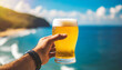 man's hand holding fresh pint of beer against blurred ocean background