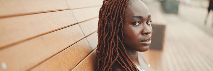 Wall Mural - Panorama of gorgeous woman with African braids wearing in top and trousers is resting sitting on bench an looks at camera