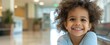 Little latino hispanic curly girl sitting in hospital or pediatric clinic on blurred light waiting room hallway background