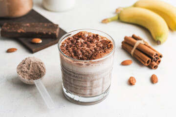 Wall Mural - Glass with chocolate protein drink, milkshake smoothie on white table with bananas, protein powder in measuring spoon, protein bar, almond nuts and cinnamon sticks