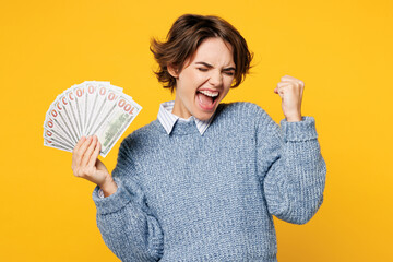 Wall Mural - Young woman wears grey knitted sweater shirt casual clothes hold in hand fan of cash money in dollar banknotes do winner gesture isolated on plain yellow background studio portrait. Lifestyle concept.