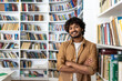 © Liubomir - A cheerful young man with curly hair stands confidently with his arms crossed in a well-stocked library. He projects a vibe of intelligence and positivity amidst a collection of books.