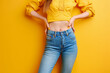 Cropped photo of model girl posing against yellow wall. Slender woman with toned stomach poses in jeans and top
