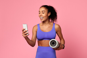 Wall Mural - Athletic African woman holds smartphone and foam roller, pink background