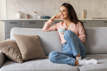 Wall Mural - Cheerful woman enjoying cup of coffee on cozy sofa