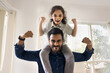© fizkes - Cheerful strong Indian dad lifting little daughter kid on shoulders, making power hands gestures, showing fists, muscles, looking at camera with martial scream, playing active games