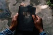 © Jouni - Black person holding a black book with the words 'VOTE 2024', pondering the right choice before an election.