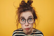 Funny cross eyed girl with messy bun hair wearing glasses and striped shirt on yellow background