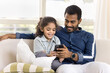 © fizkes - Happy pretty little Indian daughter kid and handsome young dad watching funny online content on smartphone, using Internet technology on mobile phone, resting on couch with gadget together