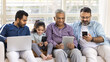 © fizkes - Online addicted Indian kid and men of four generations sitting together on sofa, using application, service, digital gadgets, holding laptop, smartphone, tablet, playing, browsing social media