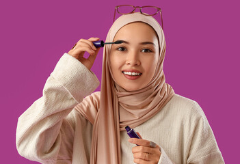 Sticker - Young Muslim woman applying mascara on purple background, closeup