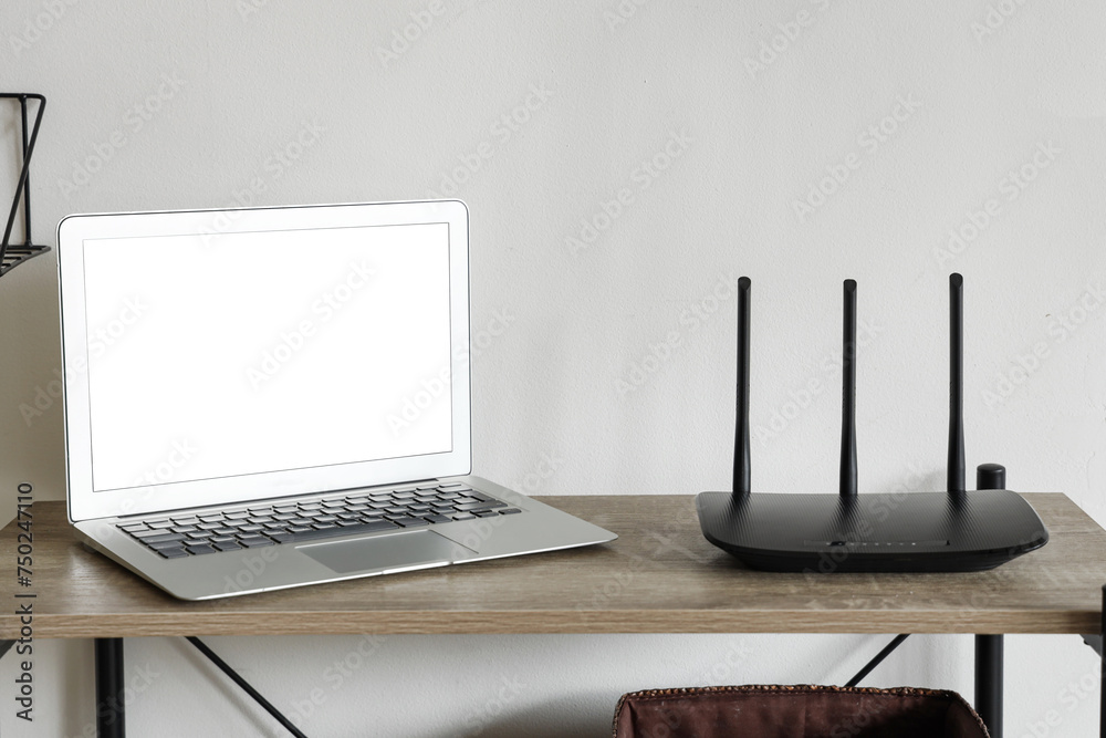 Modern wi-fi router with blank laptop on shelf near light wall