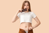 Fototapeta Panele - Young woman in tight pants eating chocolate on beige background. Weight gain concept