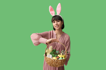 Canvas Print - Pretty young woman in bunny ears holding wicker Easter basket with cosmetics on green background