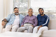 © fizkes - Happy young adult Indian grandson and son spending leisure time with older grandpa and mature father. Men of three family generations sitting on couch, looking at camera for portrait, smiling