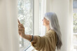 © fizkes - Happy inspired senior Latin woman opening white transparent veils, enjoying morning daylight, looking out of large window, smiling, relaxing in comfortable apartment. Side view,