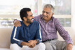 © fizkes - Cheerful elderly Indian granddad and happy young adult grandson talking on home couch, hugging, laughing, having fun, enjoying friendship, support, family communication