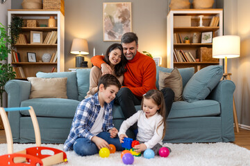 Poster - Cute kids playing while parents relaxing sofa at home together, smiling active boy entertaining with toy car near his sister on floor, happy family spending time together in living room on weekend