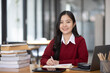 © SOMKID - Happy young asian woman wearing earphones and using laptop computer at desk in office, Female student with stack of books and laptop.