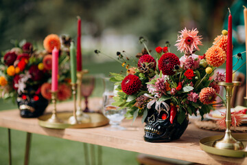 Wall Mural - Variegated multi-colored flower arrangements in vases in the shape of a skull, Mexican style in the decor. On the festive table in the wedding banquet area, compositions of flowers and greenery, candl