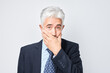 Mature businessman covering mouth with hand, speechless secret keeping concept on grey background.
