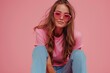 An attractive young woman in sunglasses sits on a skateboard against a pink background