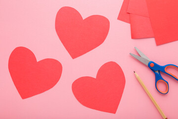 Wall Mural - Red heart shapes, scissors, pencil and application paper on light pink table background. Pastel color. Closeup. Making decoration elements. Top down view.